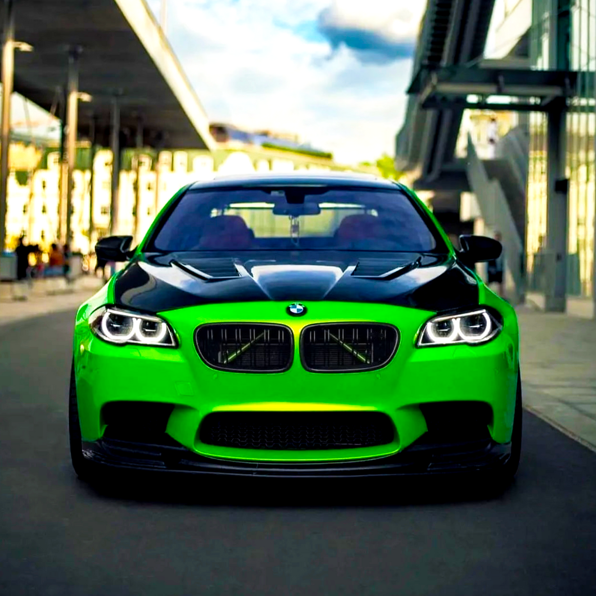 Green BMW car, parked on urban street, features black hood and prominent front grille with illuminated headlights; modern architectural structures and blue sky in the background.