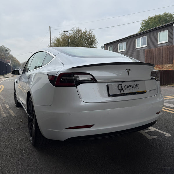 A white Tesla car is parked on a street near residential buildings. Text reads "CARBON ACCENTS," and "TESLA.COM" on the license plate frame. Overcast sky overhead.