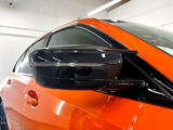 A sleek black side mirror protrudes from an orange car, reflecting interior lights in a showroom with white walls and linear overhead lighting.