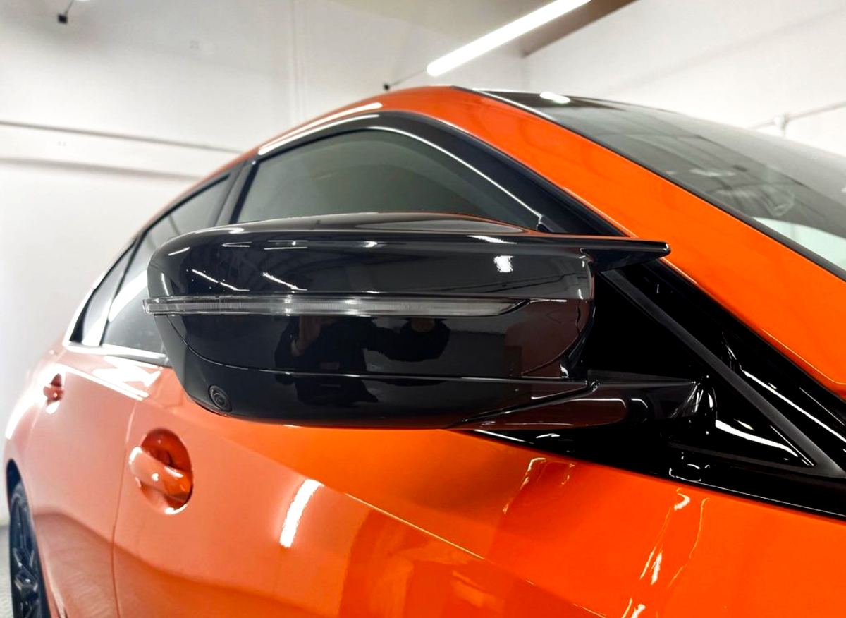A sleek black side mirror protrudes from an orange car, reflecting interior lights in a showroom with white walls and linear overhead lighting.