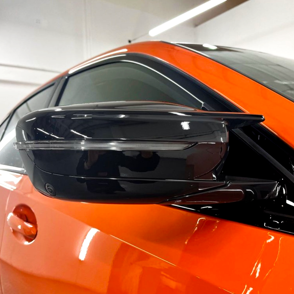 A sleek black side mirror protrudes from an orange car, reflecting interior lights in a showroom with white walls and linear overhead lighting.