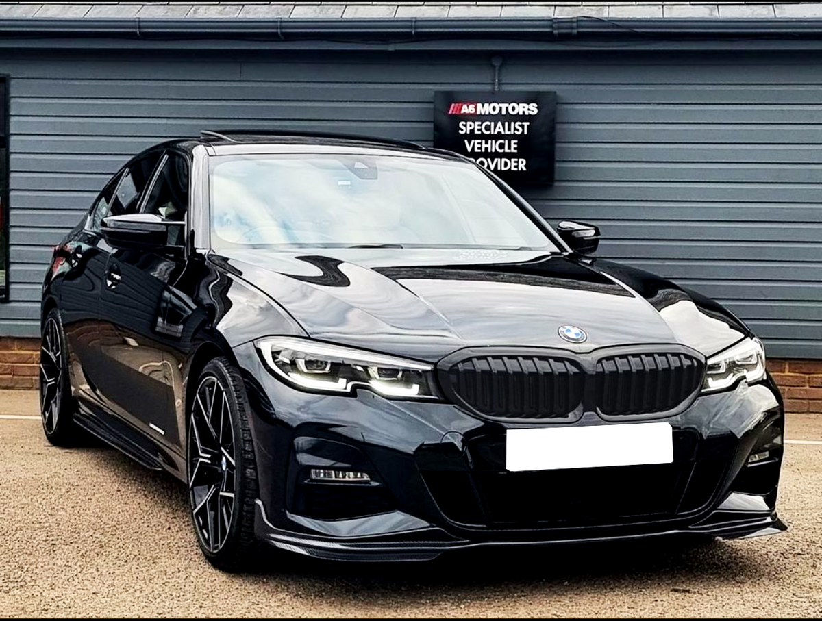A black sedan, parked in front of a gray building, features sleek design elements. The sign reads "A6 Motors Specialist Vehicle Provider." The environment appears to be a dealership.
