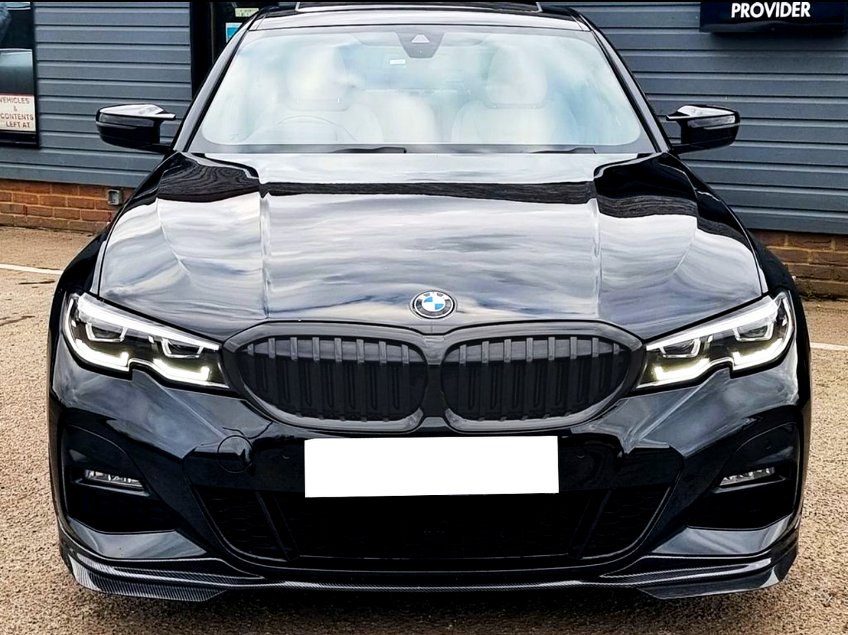 A black BMW car is parked, displaying its front grille and headlights. It is situated in a lot with a building in the background displaying the text "PROVIDER."