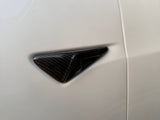 A carbon fiber car door handle resides on a smooth, light-colored vehicle door, surrounded by softly blurred ambient light, enhancing its sleek and modern design.