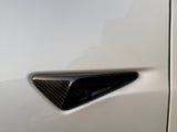 A carbon fiber side camera, embedded in a sleek, white car's body, is situated near the edge, beneath the side mirror. The setting is outdoors with soft lighting.