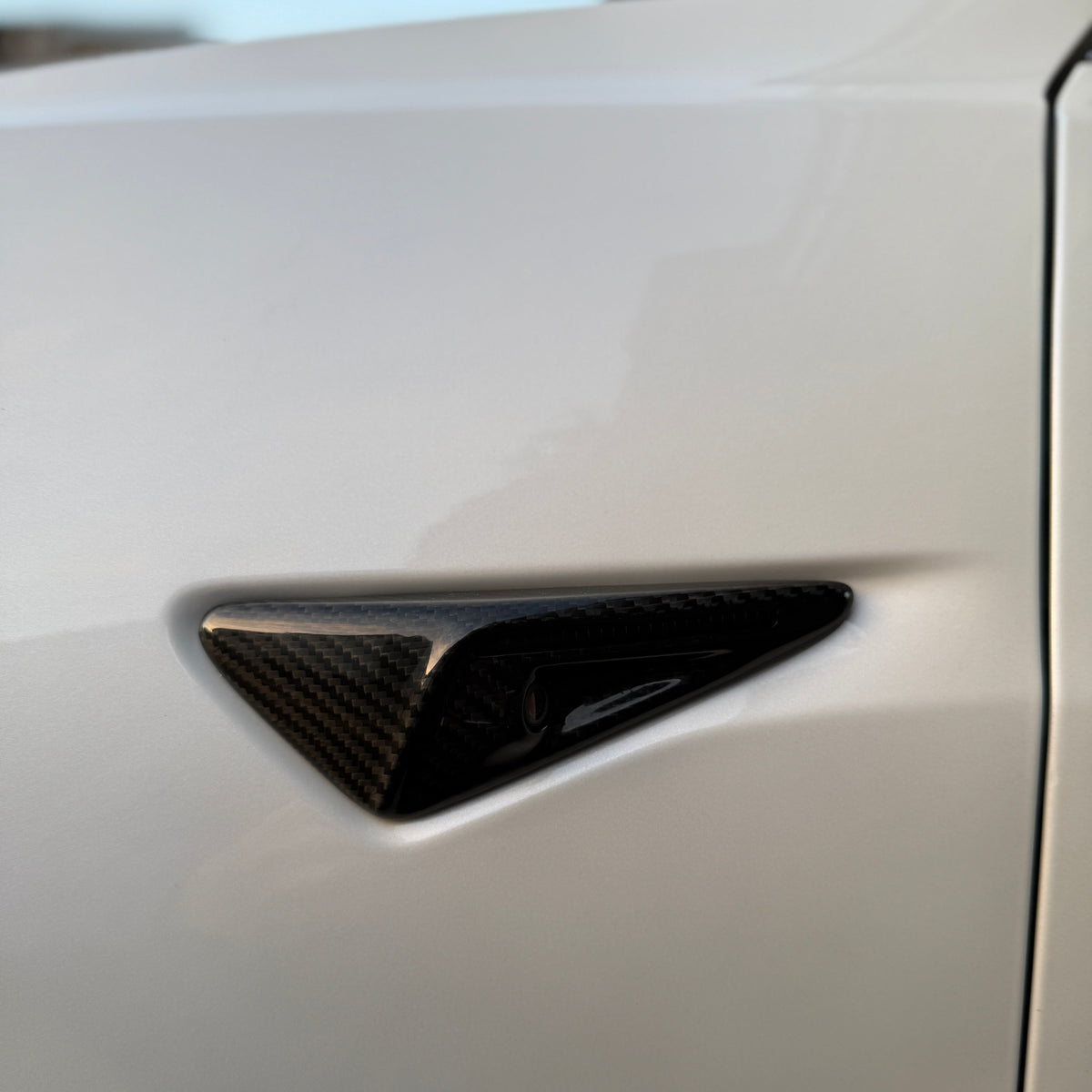 A carbon fiber side camera, embedded in a sleek, white car's body, is situated near the edge, beneath the side mirror. The setting is outdoors with soft lighting.