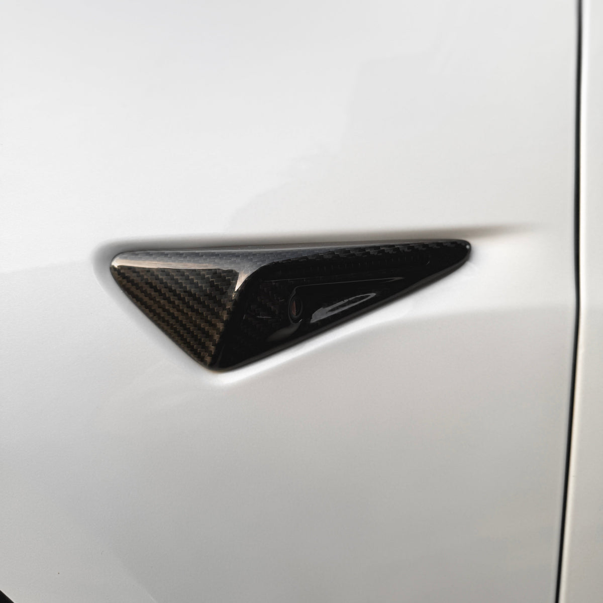 Carbon fiber trim piece, triangular in shape, embedded on a smooth, white vehicle surface. The trim adds a sleek, modern accent to the car's exterior design.