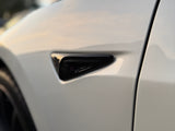 A sleek front fender vent with a glossy black finish is mounted on a shiny white car, reflecting soft ambient light, set outdoors.