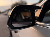 A car side mirror reflects a blurred suburban street at sunset. The vehicle's white door is partially visible, while soft light creates a warm ambiance.