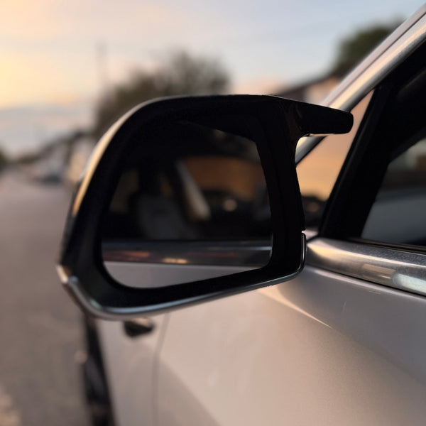 A car side mirror reflects a blurred suburban street at sunset. The vehicle's white door is partially visible, while soft light creates a warm ambiance.