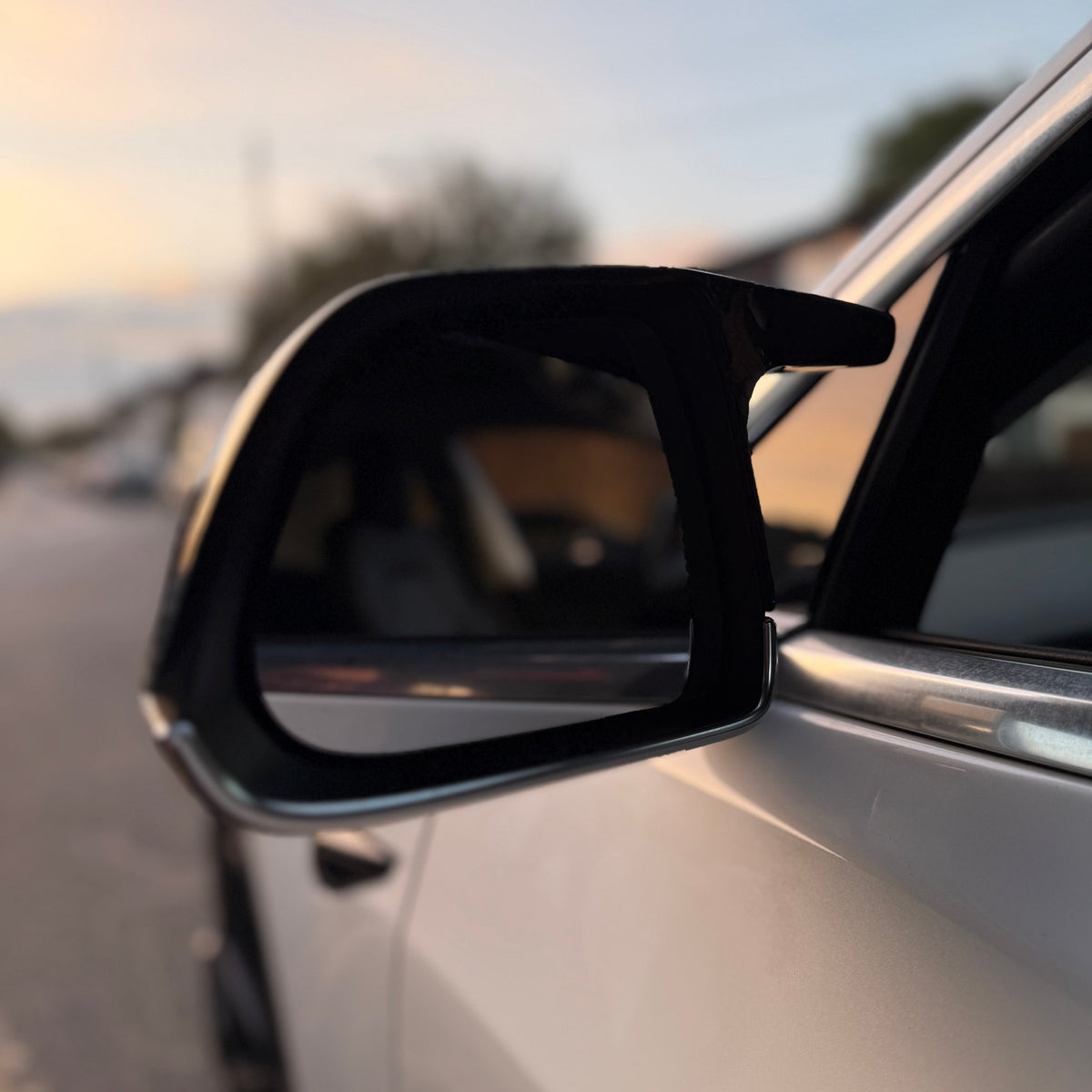 A side-view mirror reflects blurred surroundings on a parked vehicle during sunset, with a soft-focus background of a quiet street and distant trees.