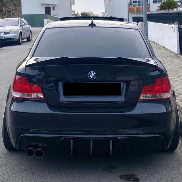 A black BMW car is parked on a residential street. The rear is prominently featured, displaying tinted windows and dual exhaust pipes. Nearby is another parked vehicle and houses.