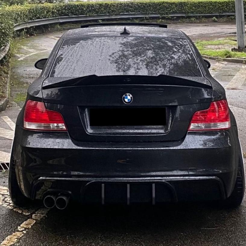 Car, stationary in a wet, winding road environment; it's black with tinted windows, dual exhausts, and a BMW logo. Surrounding context includes greenery and guardrails.