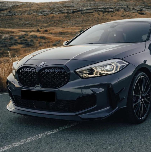 A sleek gray car with illuminated headlights is parked on a road, surrounded by a rugged, grassy landscape. The front grille is distinctive, and the wheels feature intricate designs.