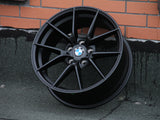 A black alloy wheel with the BMW logo rests against a brick wall. The wheel has multi-spoke design and 