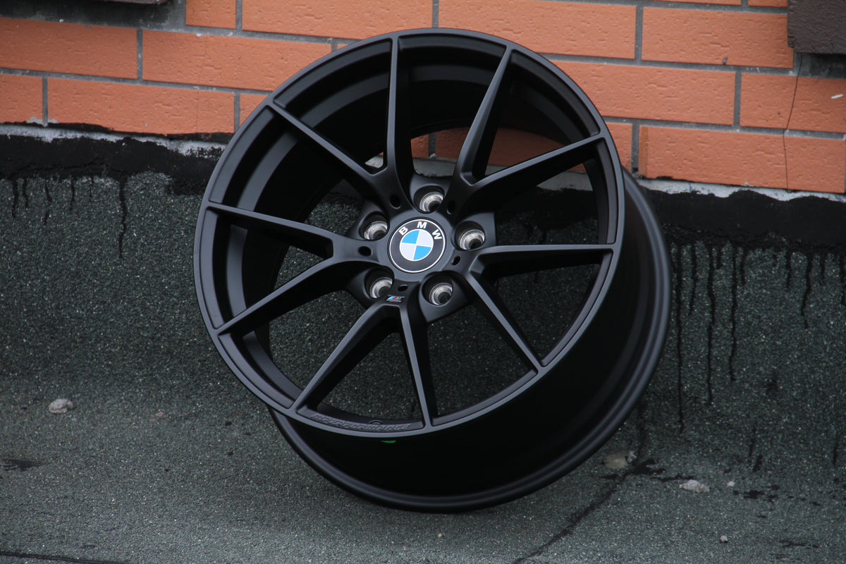 A black alloy wheel with the BMW logo rests against a brick wall. The wheel has multi-spoke design and "B M W" is visible on the center cap.