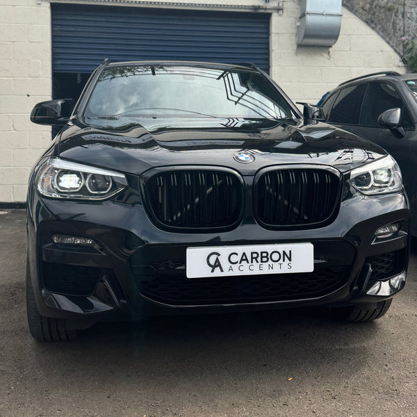 A black BMW car is parked, facing forward. The license plate reads "CARBON ACCENTS." It is situated in a lot with a blue garage door in the background.
