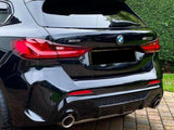 A shiny black BMW X5M parked on a brick driveway, surrounded by a brick wall and trimmed hedge, with visible red taillights and dual exhaust pipes.