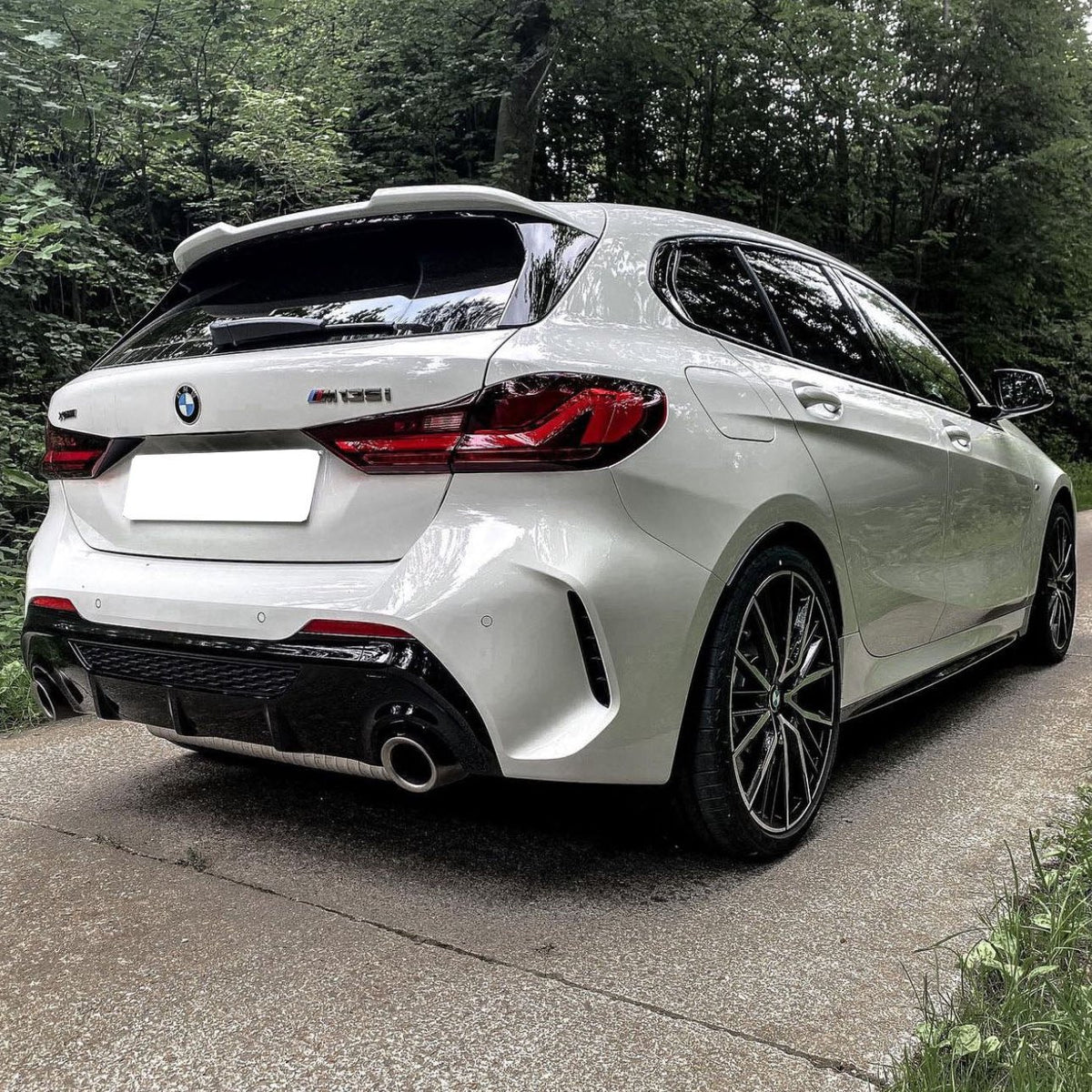 A white BMW hatchback car is parked on a narrow road surrounded by dense green forest. The model number "M135i" is visible on the rear.