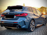 A blue BMW M135i car stands parked on a paved surface, with dual exhausts visible. In the background, trees and a setting sun create a warm glow.