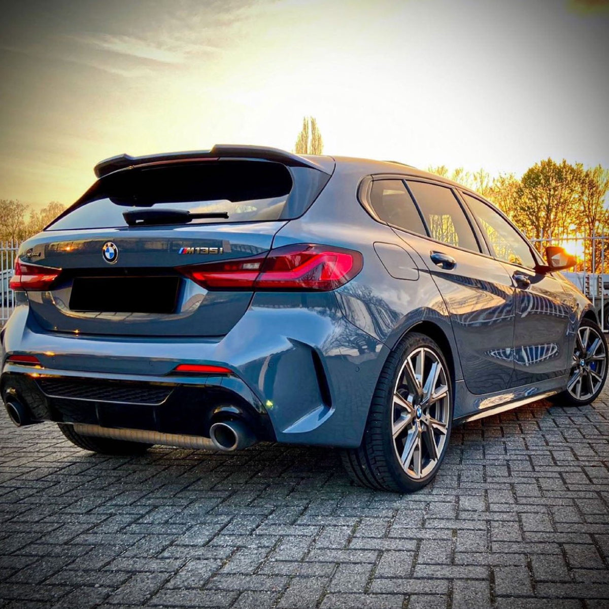 A blue BMW M135i car stands parked on a paved surface, with dual exhausts visible. In the background, trees and a setting sun create a warm glow.