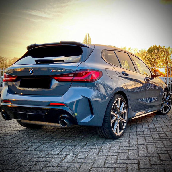A blue BMW M135i car stands parked on a paved surface, with dual exhausts visible. In the background, trees and a setting sun create a warm glow.