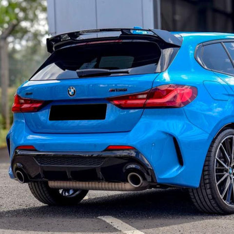 A blue BMW hatchback is parked on a street. It features dual exhaust pipes, a rear spoiler, and a visible BMW logo. Trees and a building are in the background.