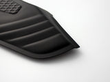 A black, ribbed car floor mat corner with a textured rectangular section rests on a smooth white surface, showing its contoured edges.