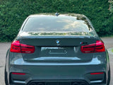 A grey BMW M3 is parked, showing the rear view with four exhaust pipes. It is situated on a paved surface, bordered by dense green hedges.