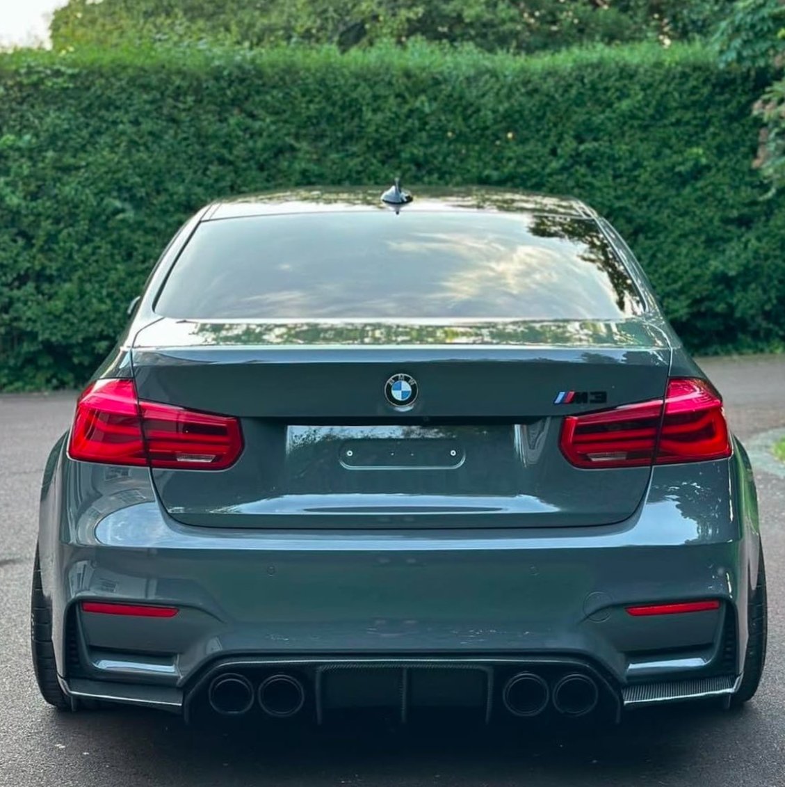 A grey BMW M3 is parked, showing the rear view with four exhaust pipes. It is situated on a paved surface, bordered by dense green hedges.