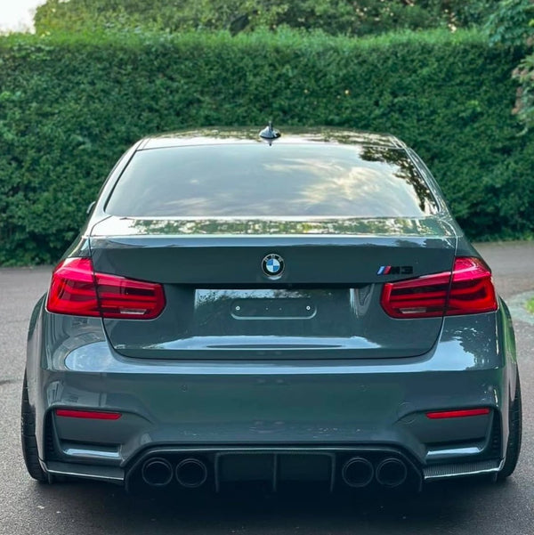 A grey BMW M3 is parked, showing the rear view with four exhaust pipes. It is situated on a paved surface, bordered by dense green hedges.