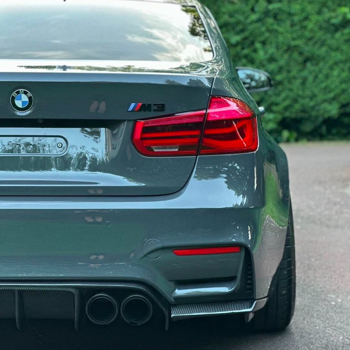 A gray BMW M3 is parked on a street. The rear features red taillights and dual exhausts, surrounded by greenery and a paved road.