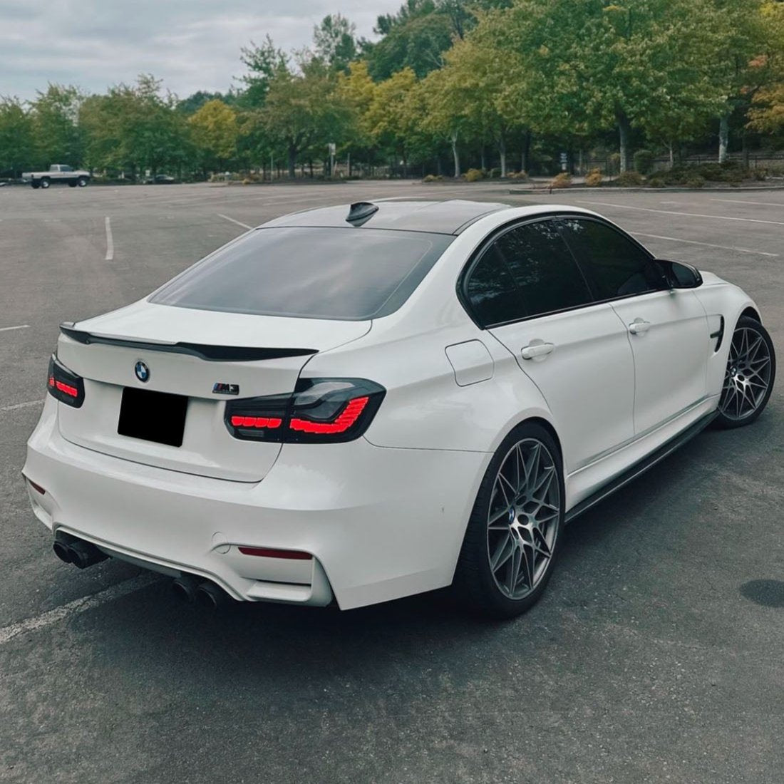 A white BMW M3 is parked in an empty lot. The car features tinted windows, dual exhausts, and distinctive alloy wheels. Surrounding the lot are lush green trees and overcast skies.