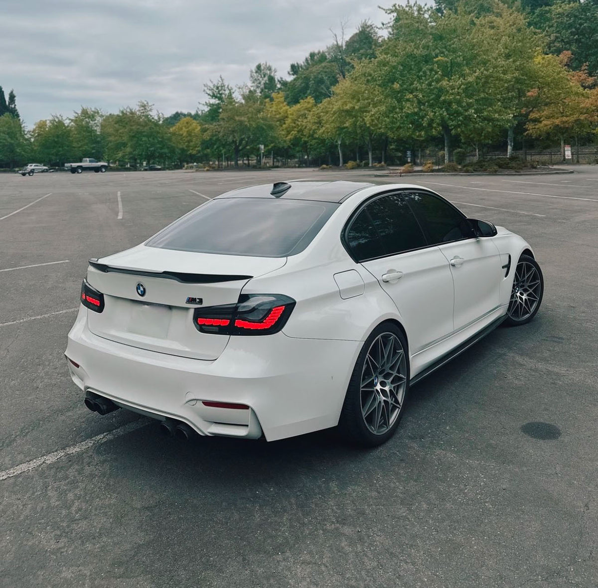 A white BMW sports sedan is parked in an empty lot. It features tinted windows, a rear spoiler, dual exhausts, and dark wheels. Green trees line the background under a cloudy sky.