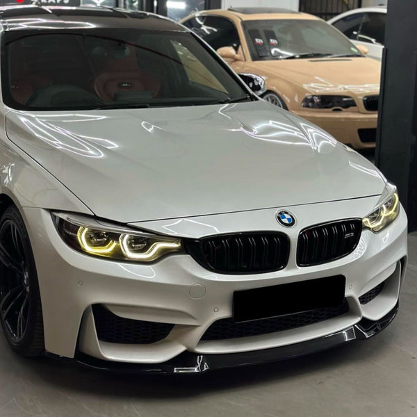A white BMW with glowing headlights is parked indoors in a showroom, with other cars visible in the background.