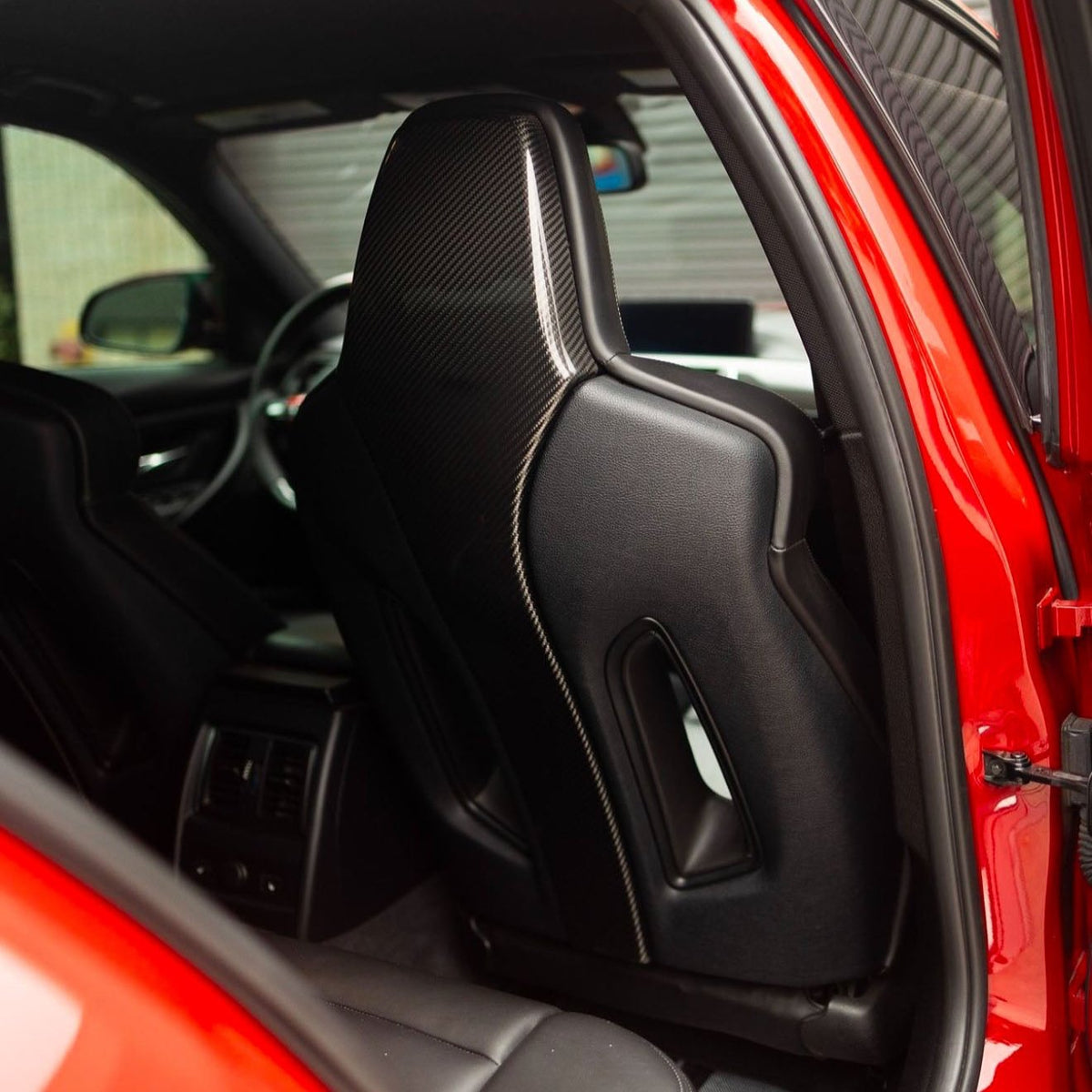 A black, sleek car seat with carbon fiber detailing is positioned inside a red vehicle, visible through the open rear door, with the steering wheel and dashboard in the background.