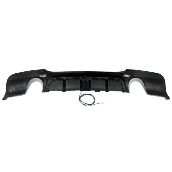 A black car bumper diffuser with integrated exhaust cutouts sits on a white background. A coiled black wire with exposed ends lies beneath it.