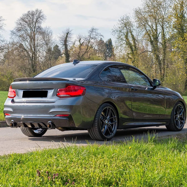 A sleek gray sports car is parked on a road, surrounded by lush green grass and tall trees, under a partly cloudy sky.