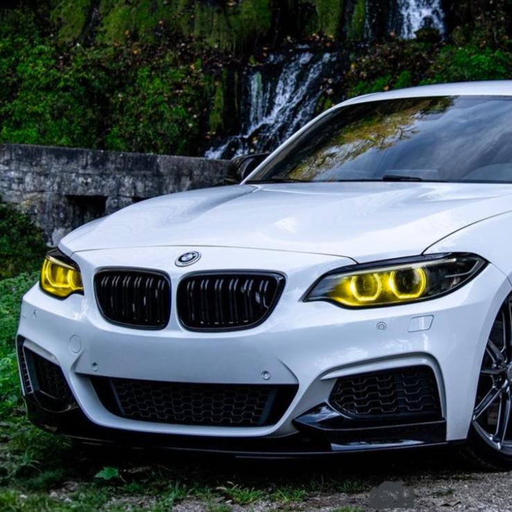 A white BMW with illuminated yellow headlights is parked on grass. Behind it, a stone wall and a small waterfall surrounded by greenery provide a scenic background.