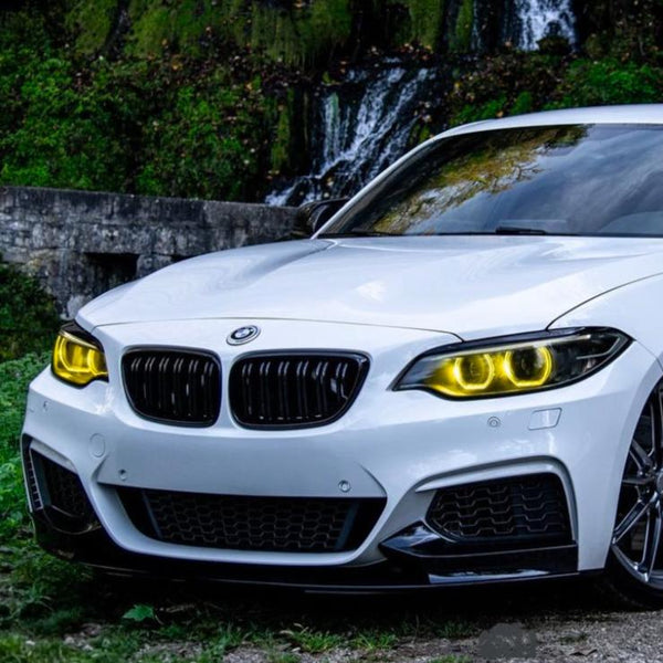 A white BMW with illuminated yellow headlights is parked on grass. Behind it, a stone wall and a small waterfall surrounded by greenery provide a scenic background.