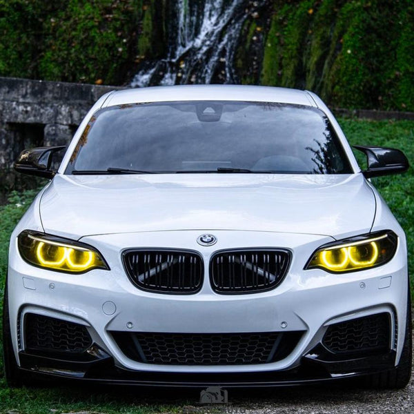 A white car with illuminated yellow headlights is parked, facing forward near a lush green area with a small waterfall in the background. No text is present.