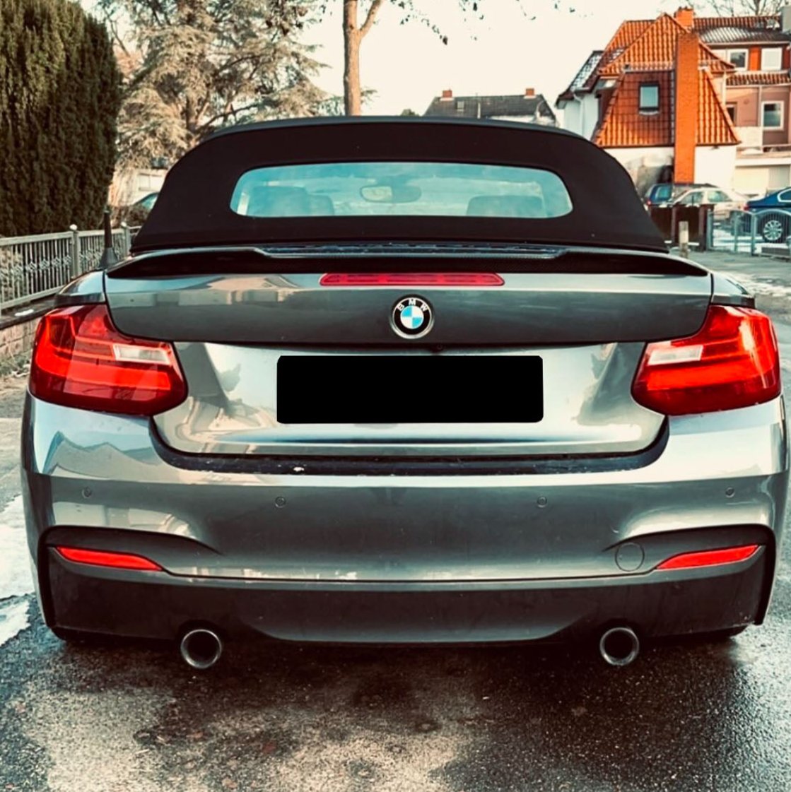 A silver BMW convertible with a black soft top is parked on a street. Surrounding it are residential buildings and trees under a clear sky.