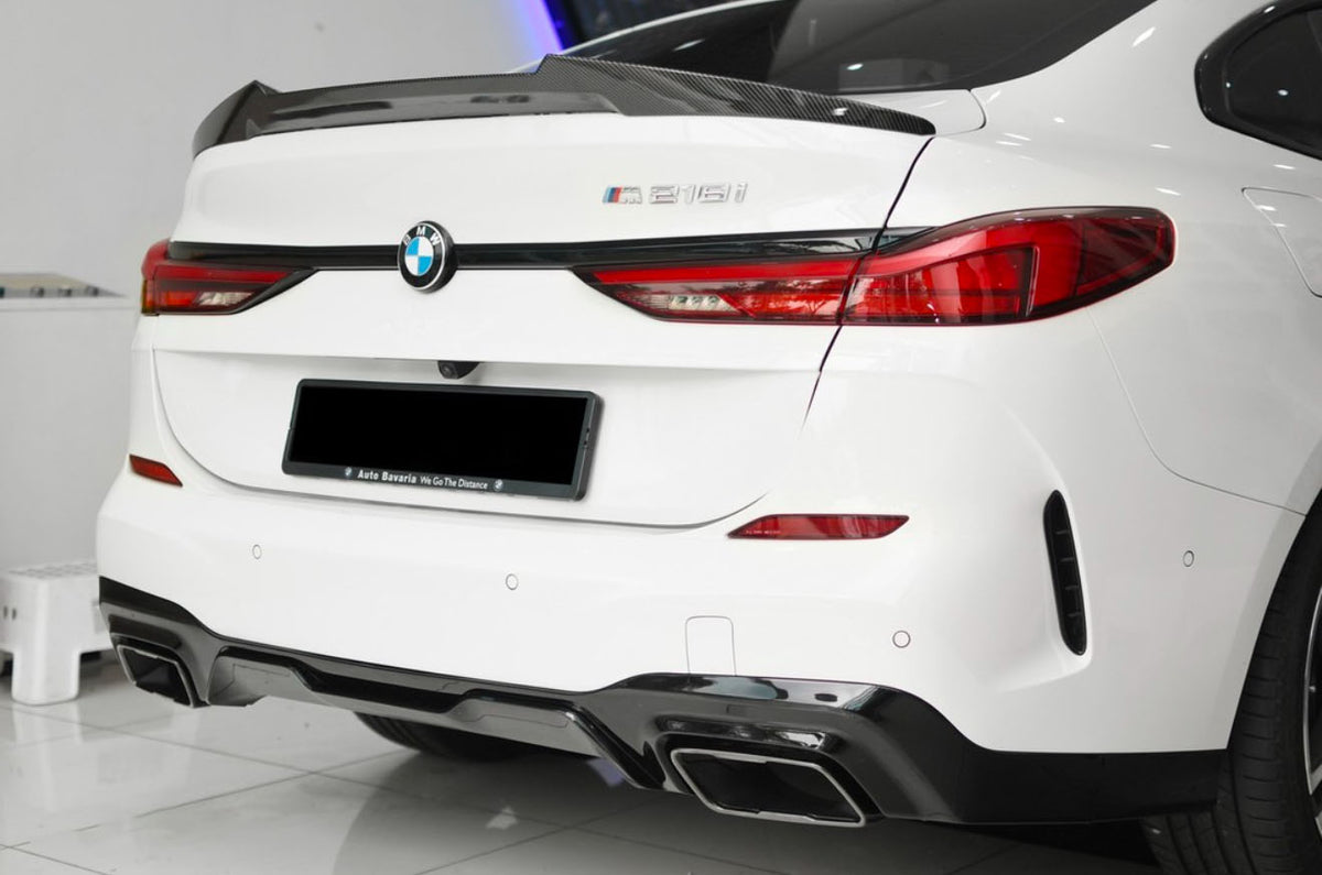 A white BMW 218i is parked indoors, featuring a carbon fiber spoiler and dual exhausts. Visible text reads "Auto Bavaria We Go The Distance" on the license plate frame.