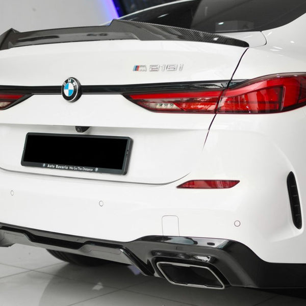 A white BMW 218i is parked indoors, featuring a carbon fiber spoiler and dual exhausts. Visible text reads "Auto Bavaria We Go The Distance" on the license plate frame.