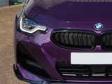 A purple car's front view, featuring a prominent grille and headlight, parked on a textured brick surface with a green bottle visible in the background corner.