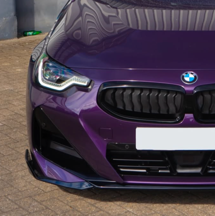 A purple car's front view, featuring a prominent grille and headlight, parked on a textured brick surface with a green bottle visible in the background corner.