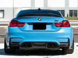 A blue BMW M4 is parked with noticeable quad exhausts and red taillights, situated in a sunny urban parking lot with a modern building backdrop.