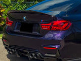 A dark purple BMW car with distinctive red taillights and dual exhausts is parked on a road, surrounded by green bushes and a clear sky reflecting on its surface.