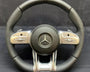 A black and silver steering wheel displays a prominent Mercedes logo at the center, surrounded by multifunctional buttons on the sides. "AIRBAG" and "AMG" are inscribed below.