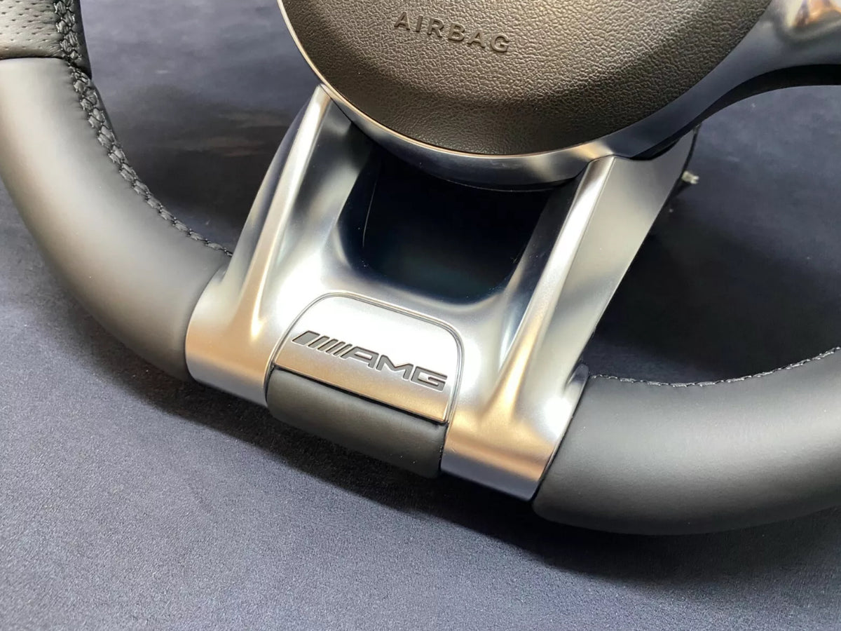 A steering wheel with "AMG" branding, featuring leather and metallic detailing. The word "AIRBAG" is embossed on the central section, set against a dark background.
